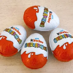 Cztery jajka Kinder Niespodzianka w pomarańczowo-białych opakowaniach na drewnianym tle.