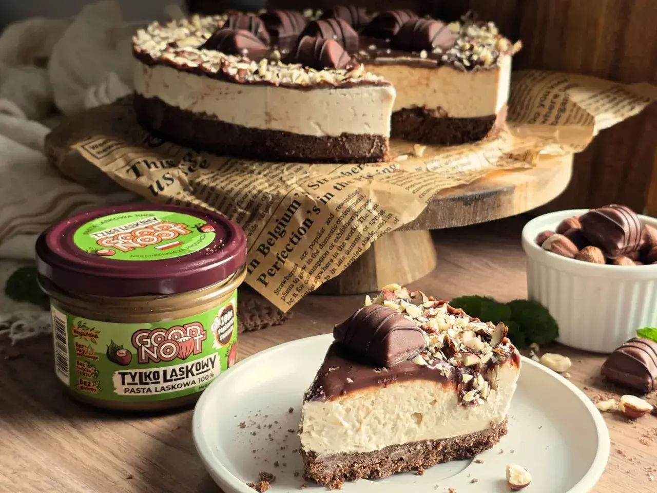 Ciasto Kinder Bueno bez pieczenia: kawałek na talerzu, obok słoik pasty orzechowej i orzechy.